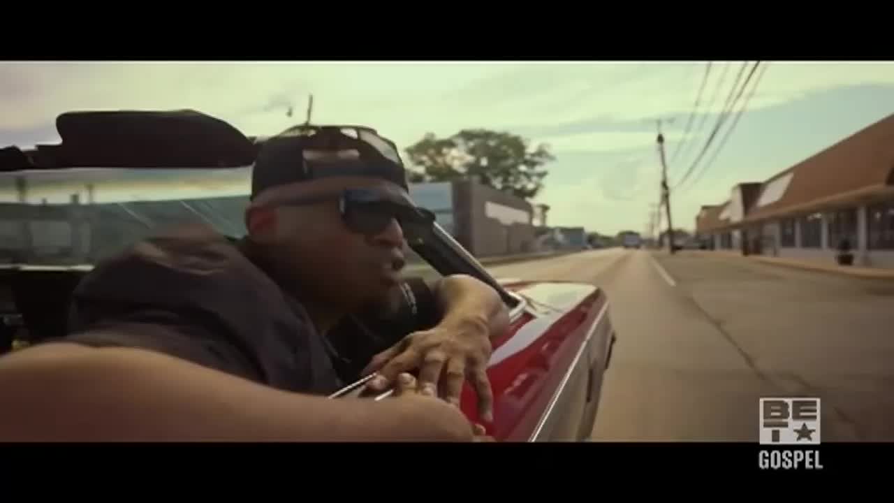 A man in a black cap and sunglasses leans out of a red convertible, looking toward the camera. The car is moving down a quiet, tree-lined street in what looks like a small American town.

