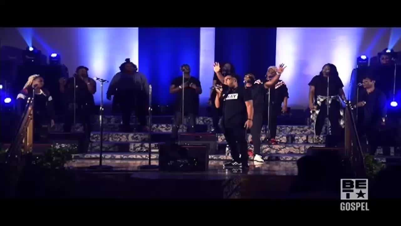 A gospel choir on a stage is singing, with some members raising their hands. The stage is lit with blue and white lights, and the BET Gospel logo is visible in the corner.
