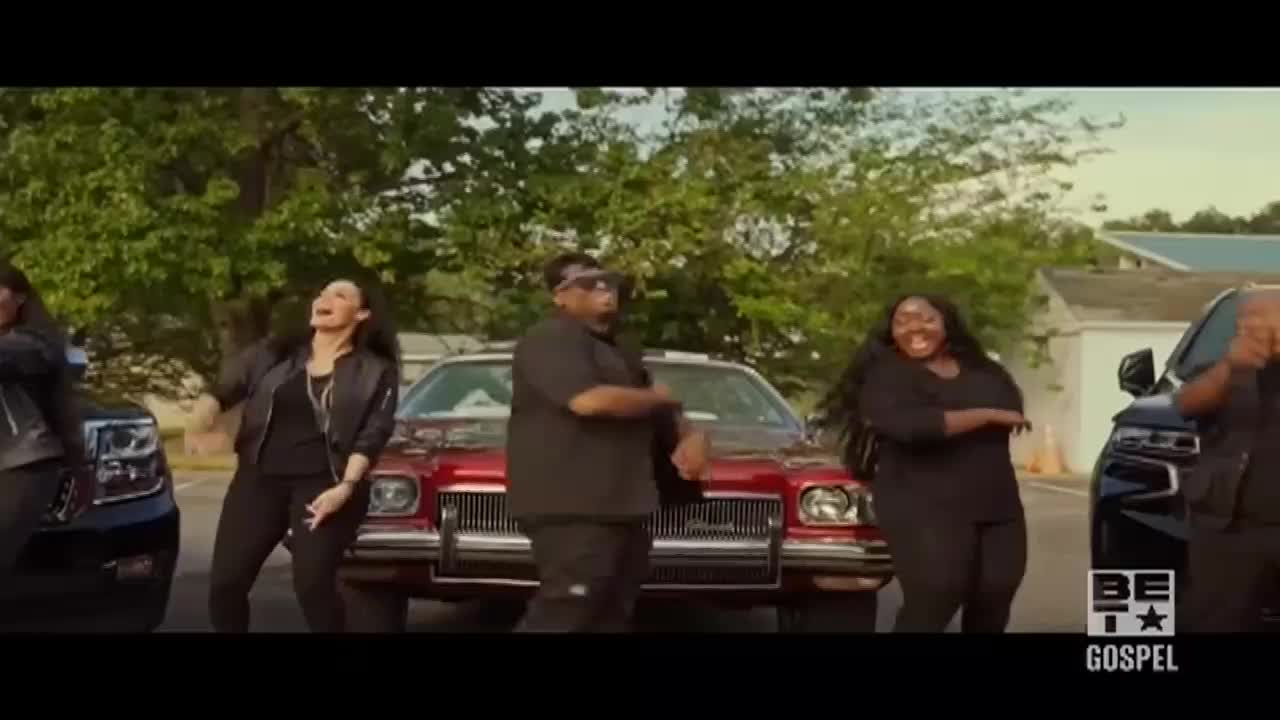 A group of people in black outfits are dancing in front of a vintage red car, with two black SUVs parked on either side. The scene is outdoors, likely in the United States, and the BET Gospel logo is visible in the corner.
