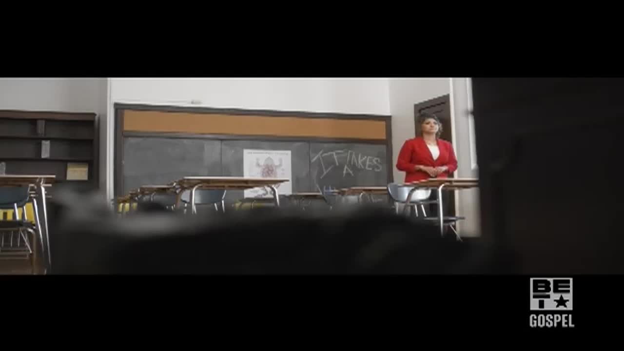 A woman in a red blazer stands behind a desk in a classroom. The chalkboard behind her has "IT TAKES" written on it.
