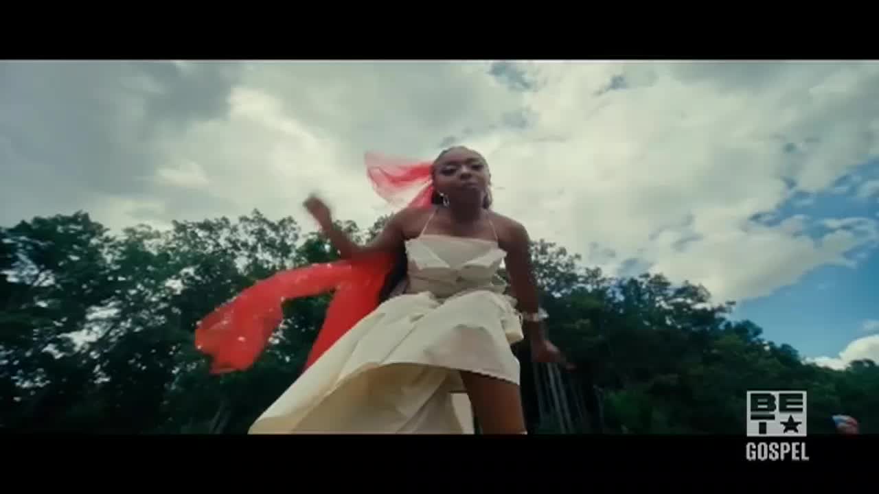 A woman in a white dress dances, a bright red scarf trailing behind her. The sky is a mix of clouds and blue as she moves, a BET Gospel logo visible in the corner.
