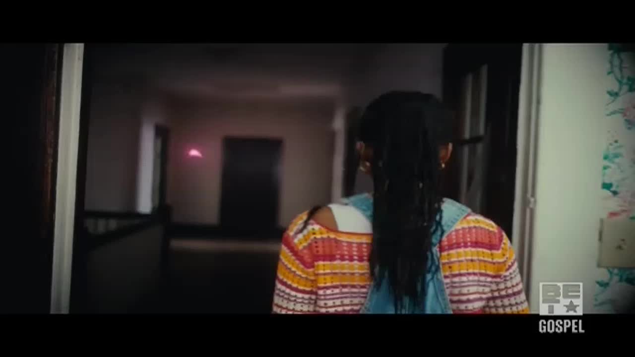 A young woman with long braids walks down a dimly lit hallway. Her colorful sweater and denim overalls stand out against the muted tones of the interior.
