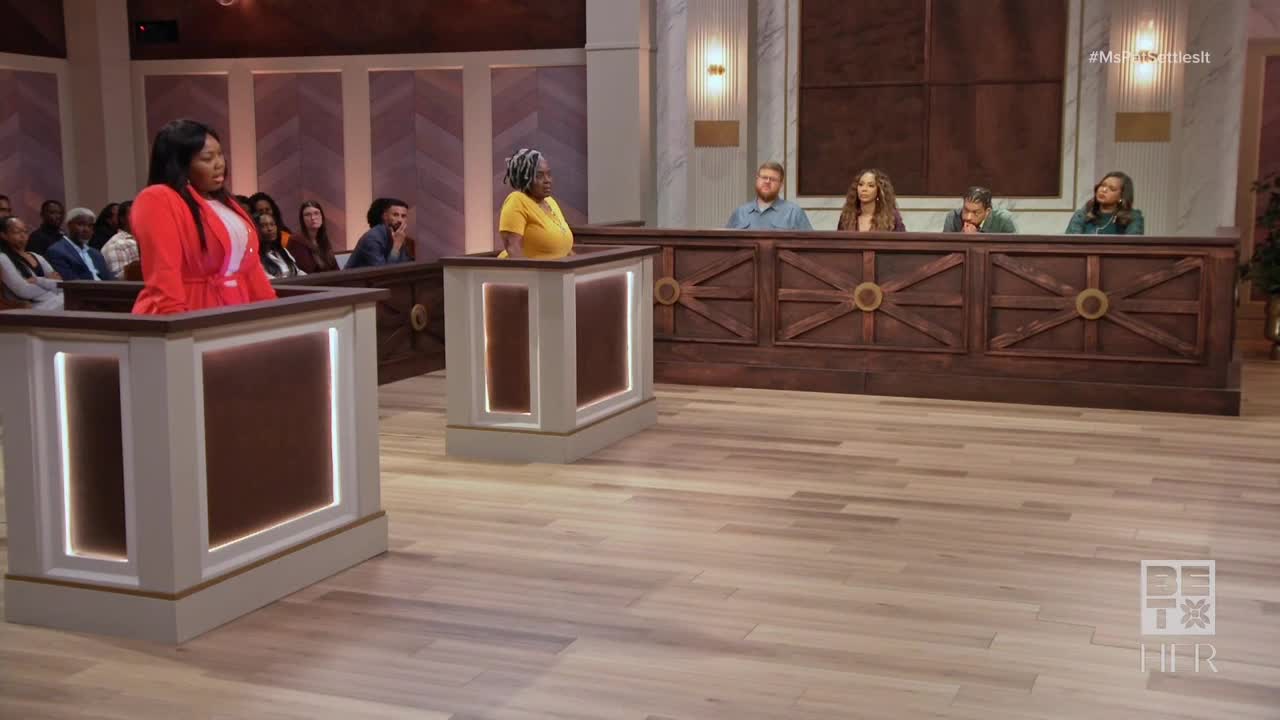 A woman in a bright red blazer stands at a podium, gesturing as she speaks. Across the room, a panel of four people sits behind a long wooden desk, listening intently.
