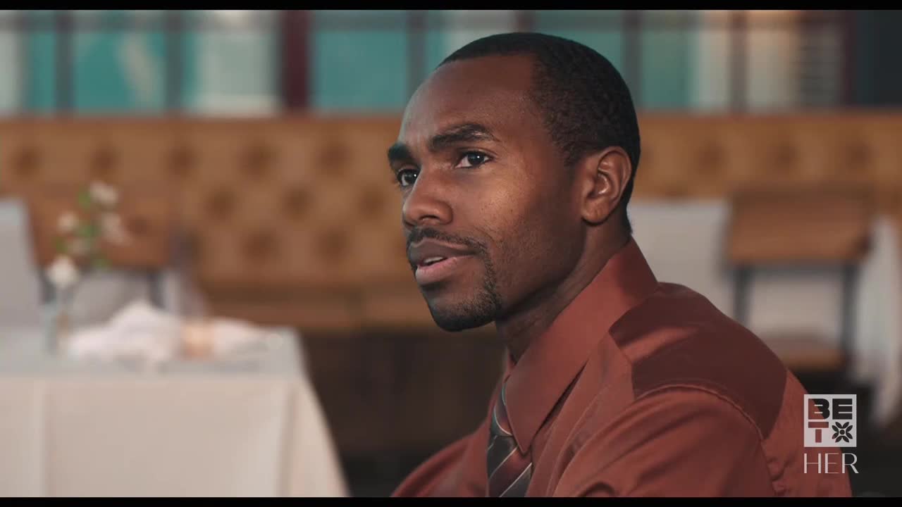 A man in a brown shirt and tie looks off to the side, seemingly listening intently. The scene appears to be set in a restaurant, possibly in the United States, as indicated by the BET Her logo.
