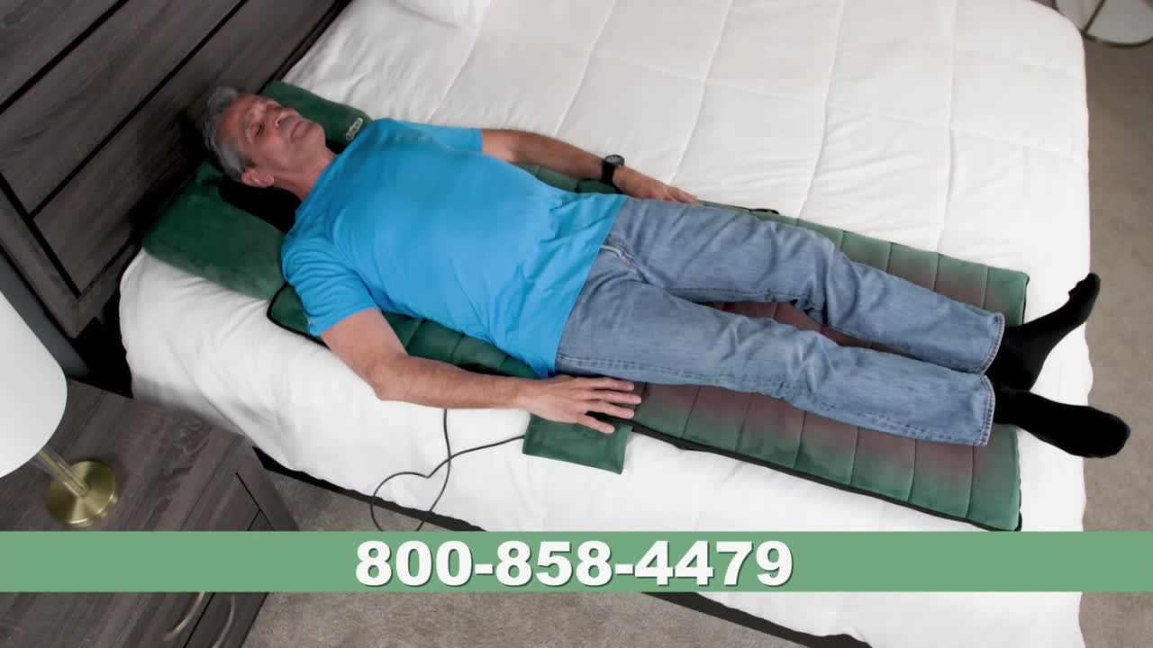 A man in a blue shirt and jeans is lying on a green and red pad on a bed. He's resting on a pillow, and a phone number is displayed at the bottom of the screen.
