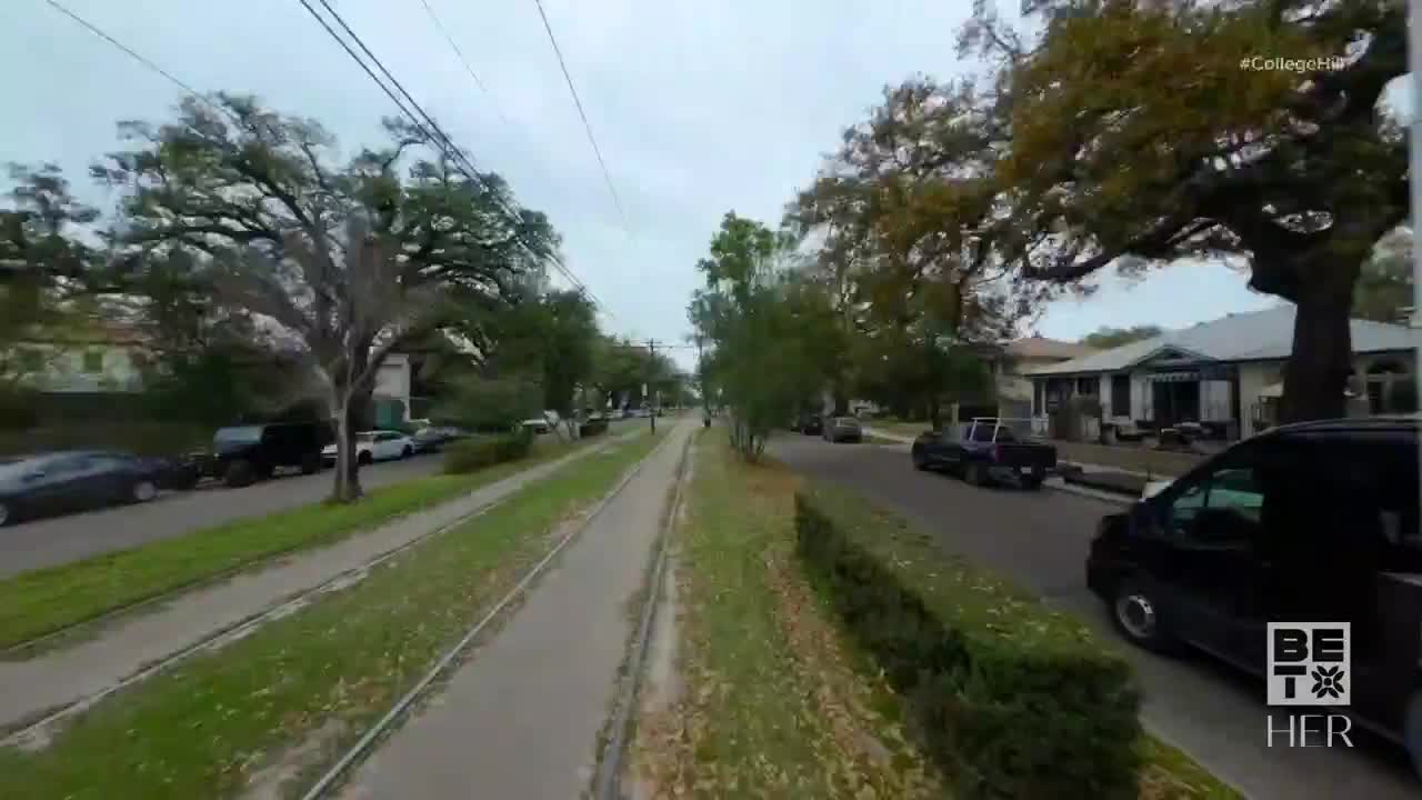 A streetcar track runs down the center of a tree-lined street. Cars and trucks are parked along the sides, and a black van is driving in the right lane.
