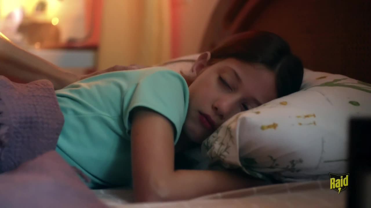 A young girl rests on a pillow, her eyes closed, as a hand gently rests on her back. The room is dimly lit, with a warm glow coming from the background.
