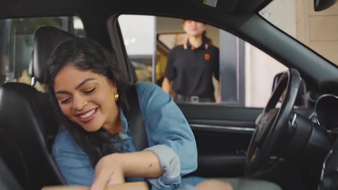 A woman in a denim jacket smiles, reaching for something in the passenger seat. A fast-food worker in a black polo shirt waits at the drive-thru window.
