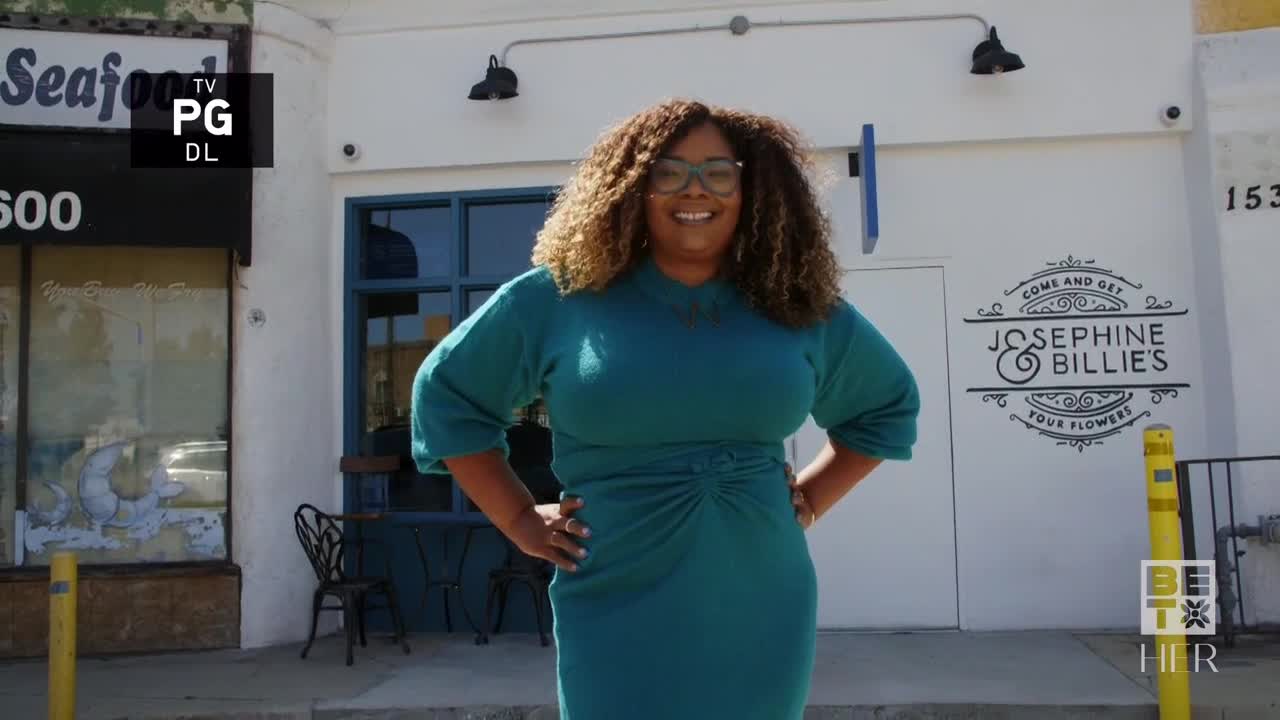 A woman in a teal dress stands in front of a business, smiling with her hands on her hips. The BET Her logo is visible in the bottom right corner.
