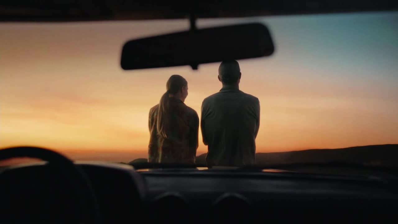 A man and a woman sit side-by-side, silhouetted against a vibrant sunset. The scene is framed by the interior of a car, suggesting a moment of reflection after a drive.

