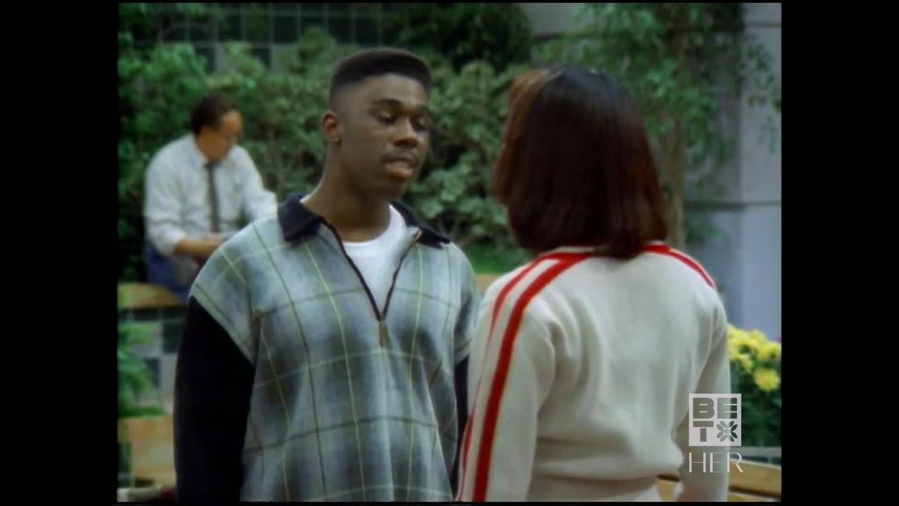 A young man in a plaid shirt speaks intently to a woman in a white jacket with red stripes. Behind them, a man sits on a bench, seemingly lost in thought.
