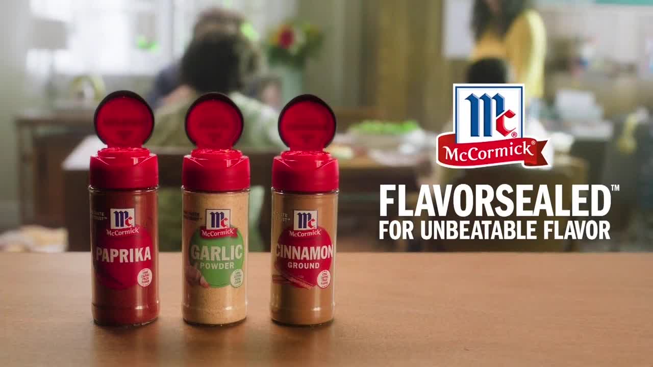 Three McCormick spice bottles with their red lids flipped open sit on a table. Behind them, a family is blurred out in the background, likely watching television on BET Her.

