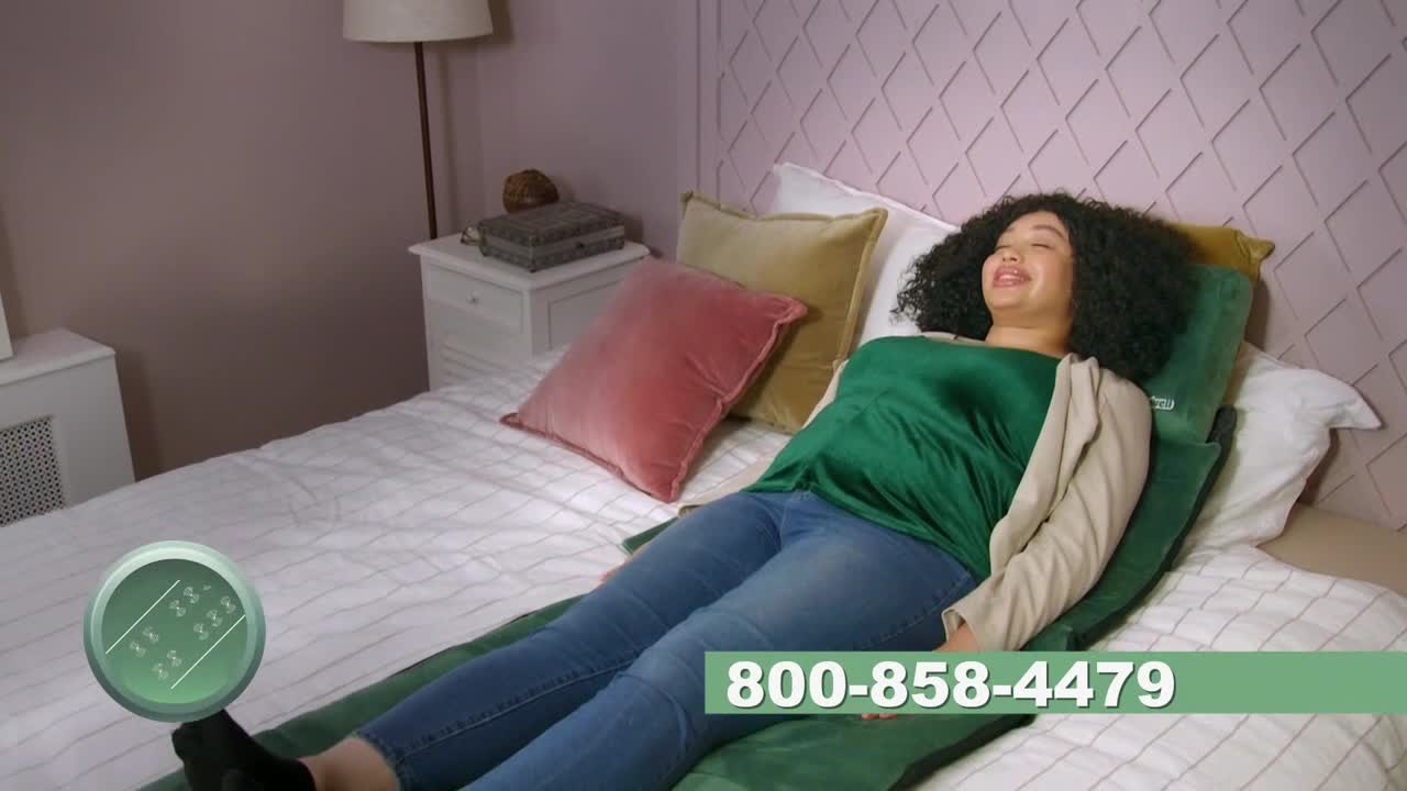 A woman reclines on a bed, resting on a green mat and smiling. A phone number for a product is displayed at the bottom of the screen.
