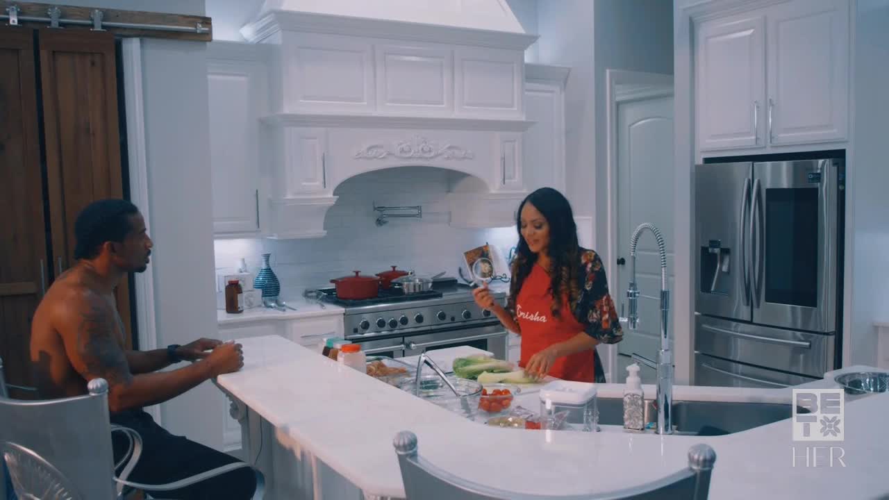In a bright kitchen, a woman in an apron is slicing vegetables. A shirtless man sits at the counter, watching.

