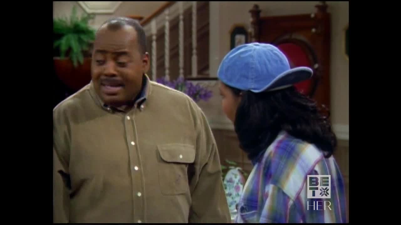 A man in a brown shirt talks animatedly, his face etched with concern. Beside him, a young woman in a blue cap and plaid shirt listens, her back to the camera.

