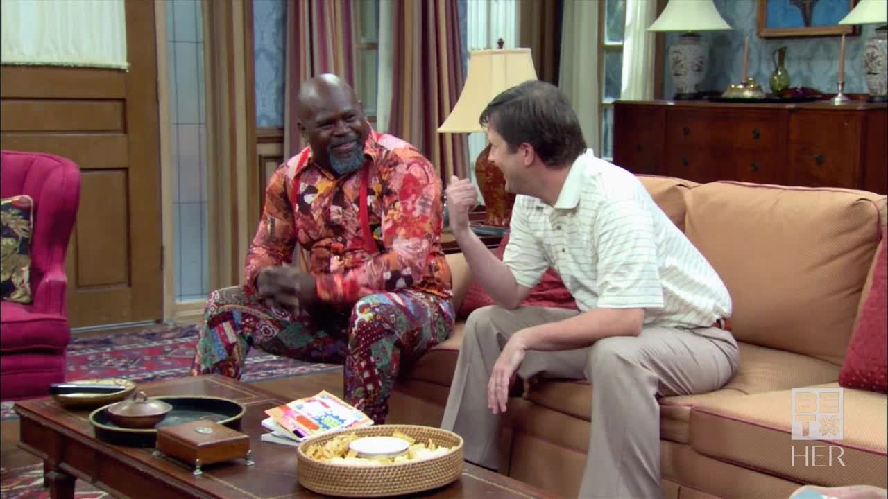 A man in a patterned shirt and matching pants grins, sitting on a couch. Beside him, another man gestures with his fist, seemingly mid-conversation.
