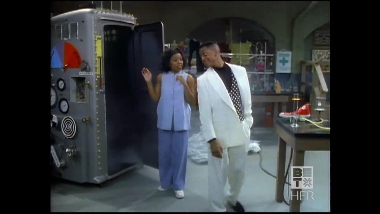 The woman in the blue outfit gestures excitedly as she walks out of a large, metallic machine. Her companion, dressed in a white suit with a black and white polka-dot tie, smiles and follows her.
