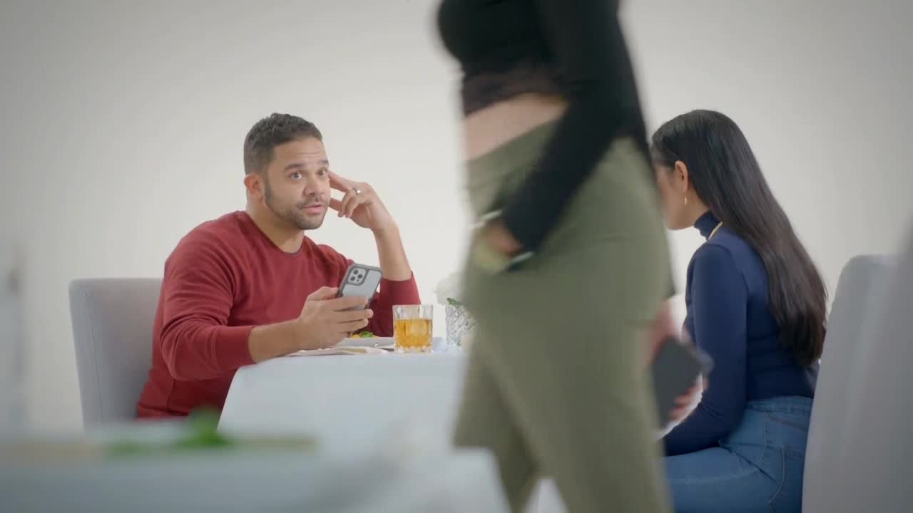 A man in a red sweater sits at a table, phone in hand, looking perplexed. A woman in a black top and green pants walks past, obscuring the other woman seated at the table.
