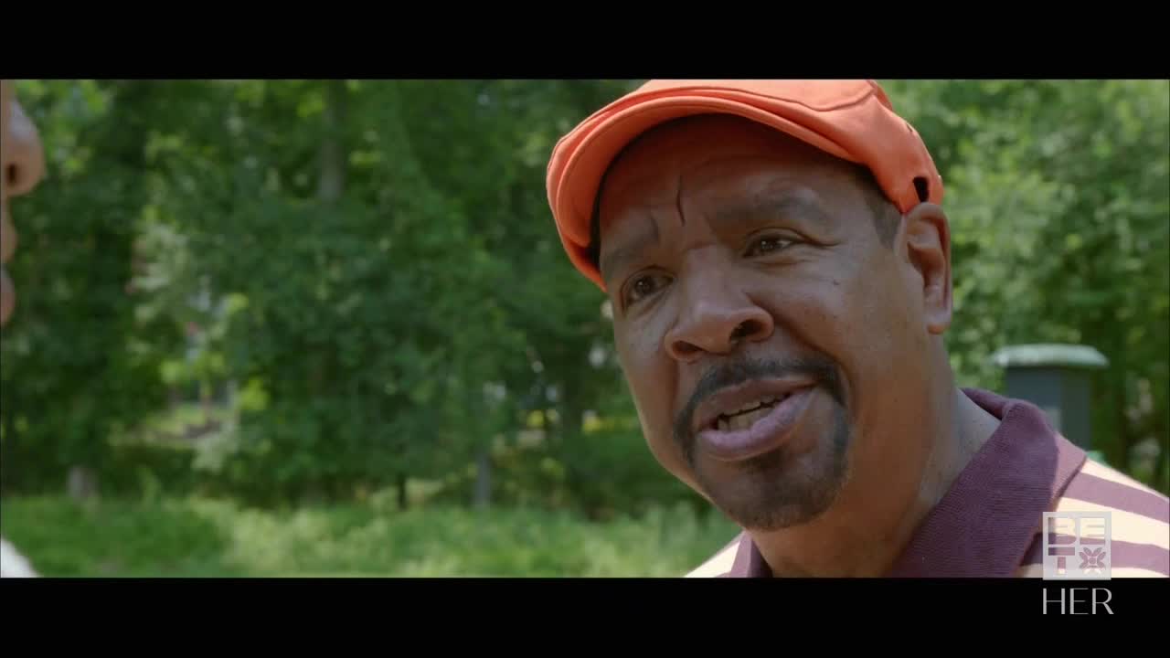 A man in an orange cap is speaking, his mouth slightly open. He's wearing a striped polo shirt, and the trees behind him are a lush green.
