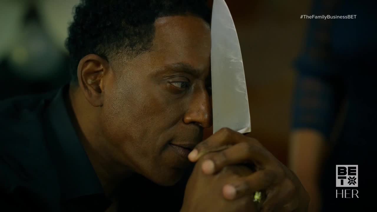 A man holds a large knife, the blade pressed against his cheek. He stares off to the side, his hands clasped together below the knife.
