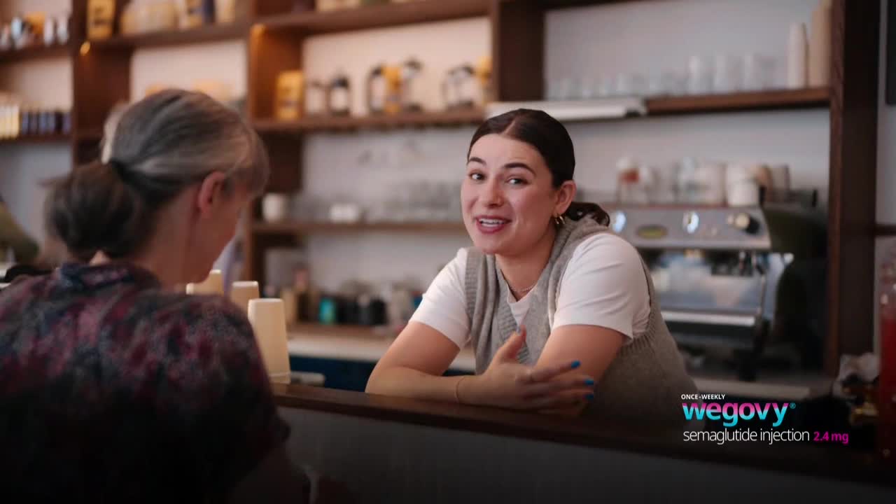 A woman behind a coffee shop counter gestures with her hands, speaking to a customer. The customer, facing away, listens intently while the barista smiles.

