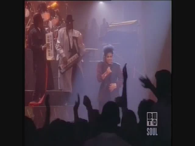 A woman in a dark suit sings into a microphone, her arms raised as the crowd cheers. Two keyboardists in suits stand behind her, bathed in stage lights.
