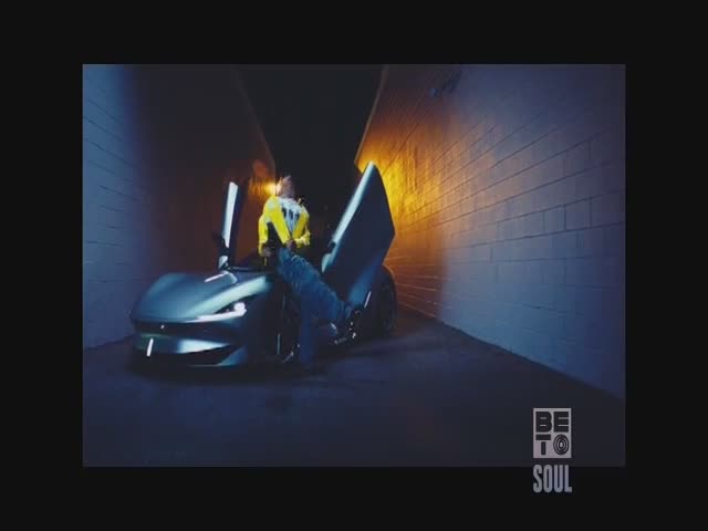 A person in a yellow jacket leans against a sleek, silver car with its doors open. The scene is dimly lit, with the BET Soul logo in the corner.
