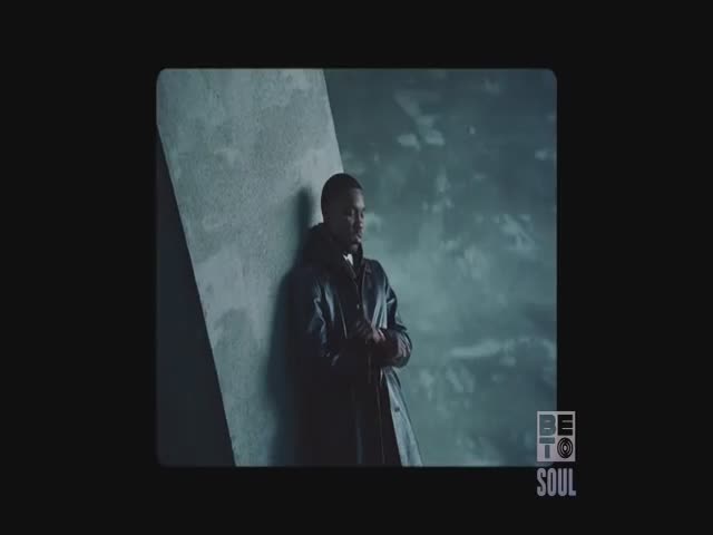 A man in a long, dark coat leans against a concrete wall. The BET Soul logo is visible in the corner, and the background is a cloudy, muted teal.
