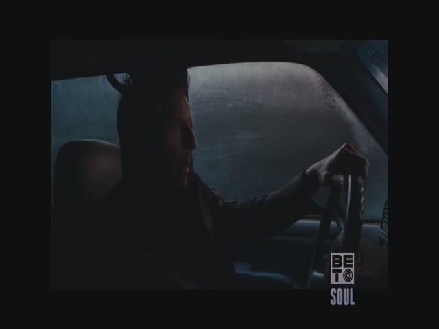 A man grips the steering wheel, his face partially shadowed. Rain streaks across the windshield, obscuring the view outside.
