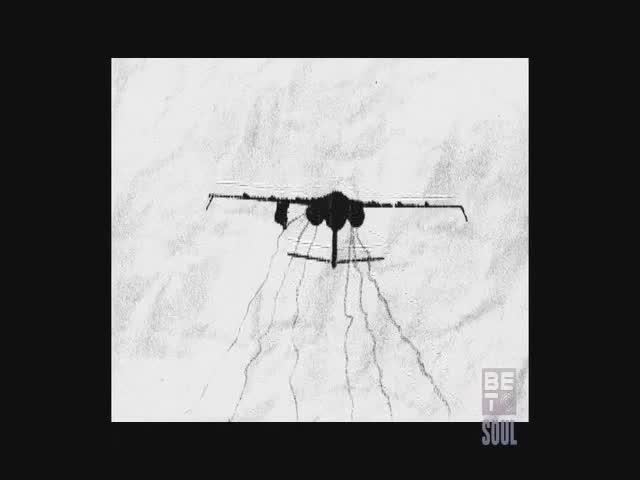 An aircraft is spraying a field, leaving trails of mist behind it. The plane's shadow stretches across the ground as it flies.
