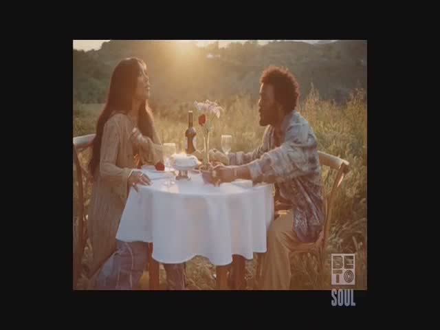A couple is seated at a table set for a meal, bathed in warm sunlight. The woman looks up, while the man gazes at her, both seemingly lost in the moment.

