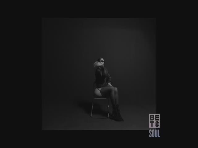 A woman in a leather jacket and boots sits in a simple metal chair. The BET Soul logo is in the corner, and the background is a stark, dark gray.
