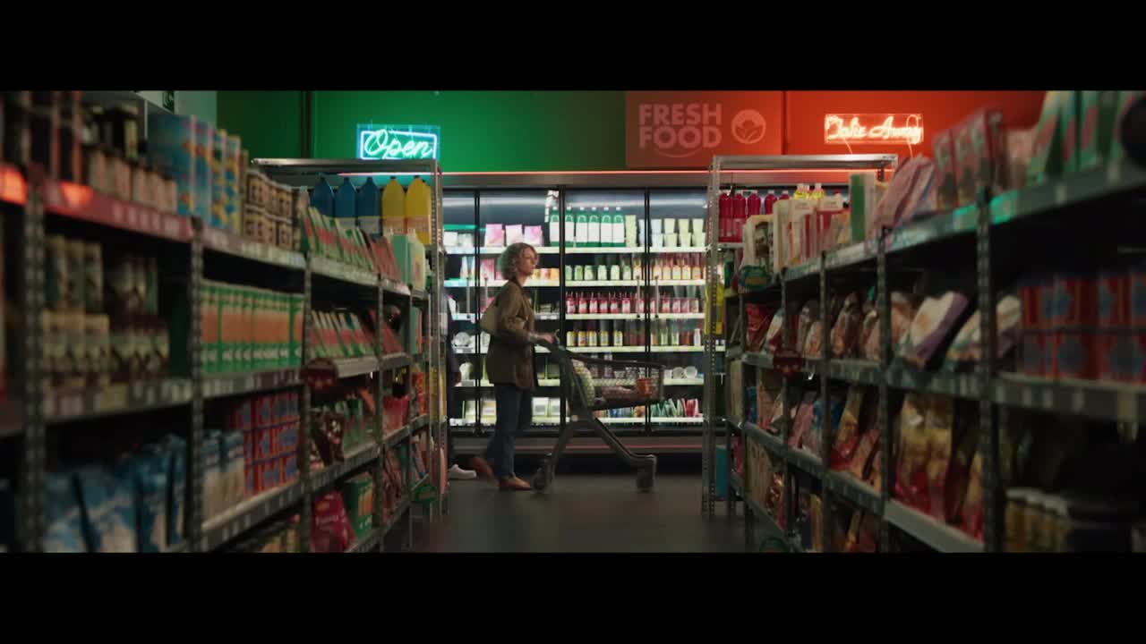 A woman pushes a shopping cart down a grocery store aisle, passing shelves stocked with various products. The bright neon signs above the refrigerated section read "Open" and "Fresh Food."
