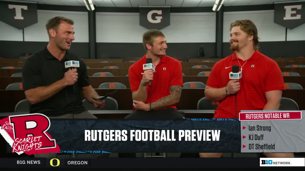 Three men sit at a table, each holding a microphone with the Big Ten Network logo. The man on the left gestures with a small tablet, while the other two, wearing red shirts, seem to be listening intently.
