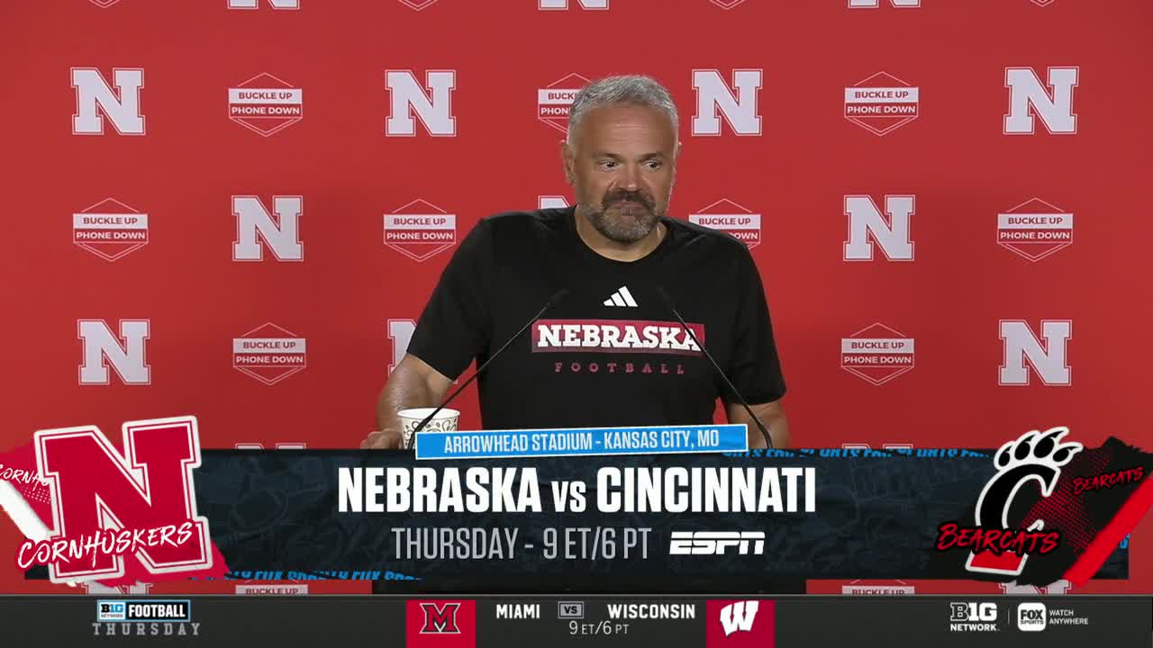 The Nebraska football coach is at a press conference, speaking into two microphones. Behind him, the Big Ten Network's backdrop displays the game's details, including the Cincinnati Bearcats.
