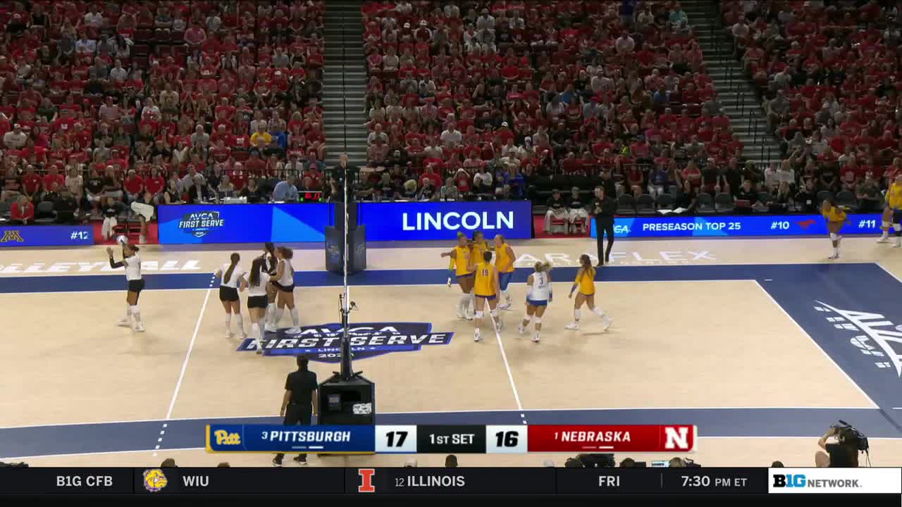 The Nebraska team in yellow jerseys huddles, celebrating a point. Across the net, the Pittsburgh team in white and black stands, regrouping after the score.

