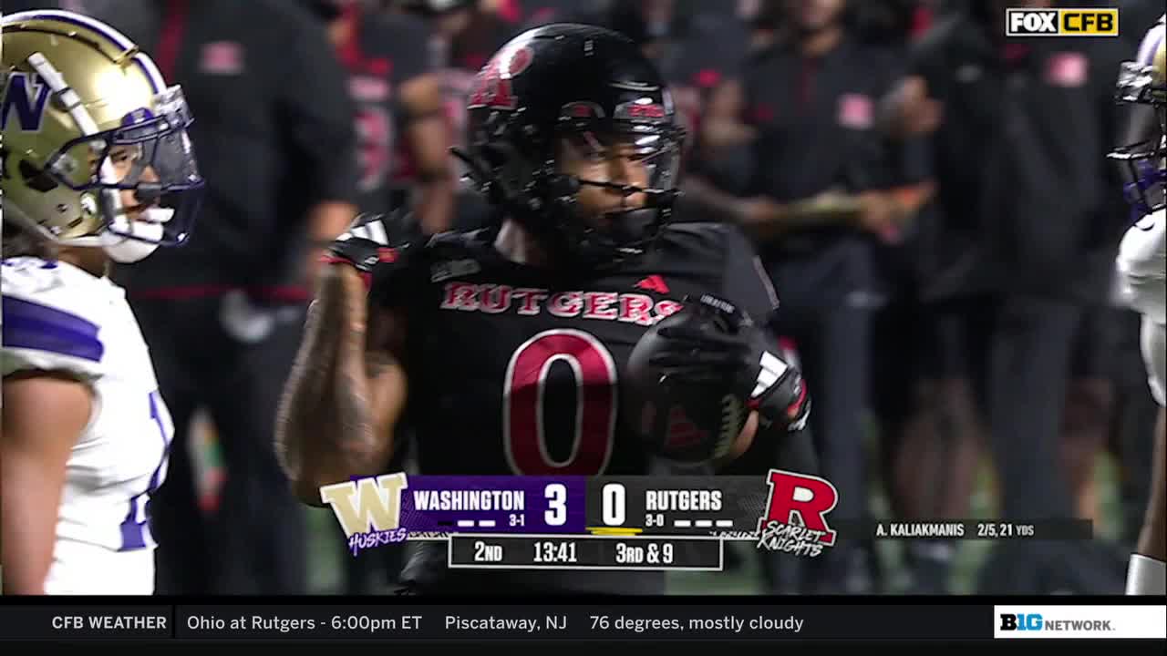 The Rutgers player, wearing a black helmet and jersey with the number zero, holds the football, looking focused. The scoreboard shows Washington leading Rutgers in the second quarter of the game, broadcast on the Big Ten Network.
