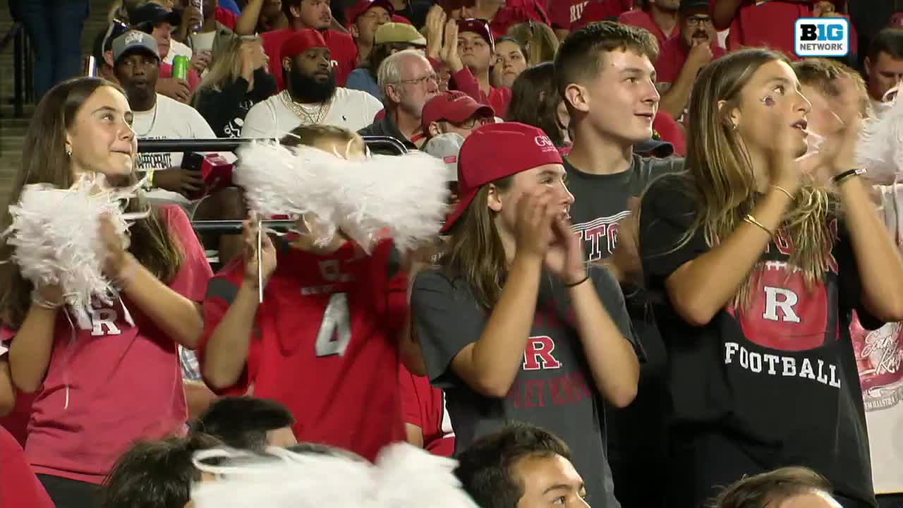 Fans in red shirts and hats cheer, clapping their hands enthusiastically. White pom-poms are raised, and the Big Ten Network logo is visible.
