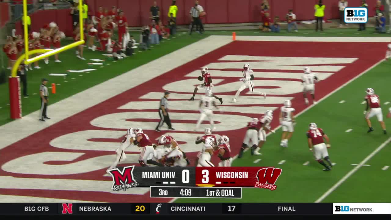 The Wisconsin Badgers are on the attack, with the ball carrier sprinting toward the end zone. A cluster of players from both teams are battling near the goal line.
