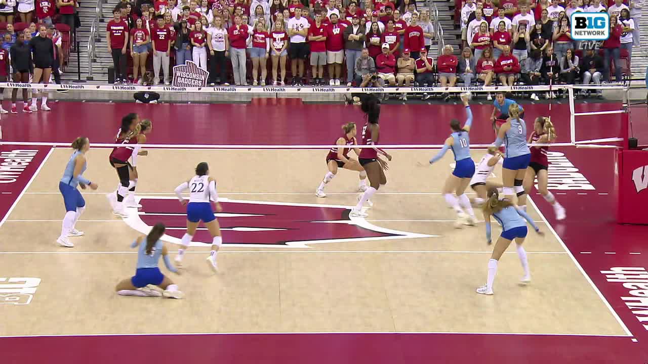 A player in a maroon uniform leaps to spike the ball over the net. A player in blue dives to the floor, trying to make the save.
