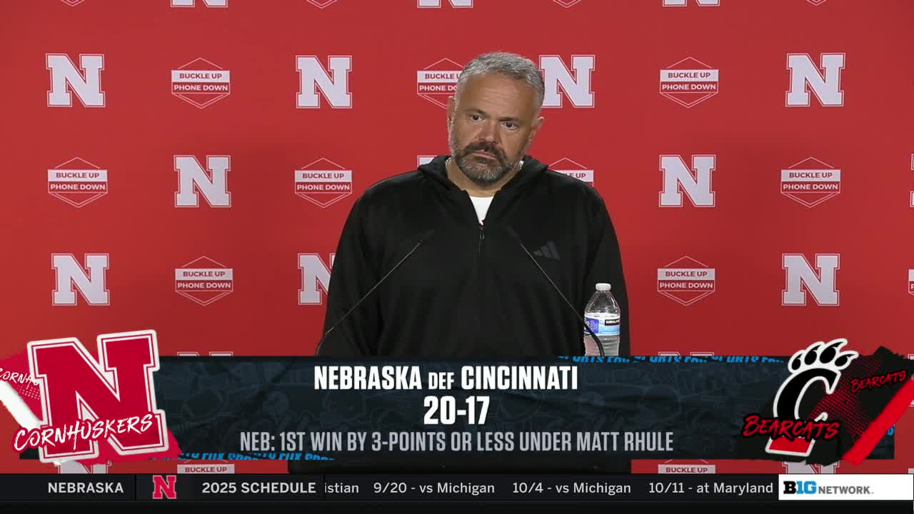 A somber-looking coach stands at a podium, a microphone in front of him, with the Big Ten Network logo visible. The scoreboard displays the Nebraska-Cincinnati score, 20-17, with the Cornhuskers victorious.
