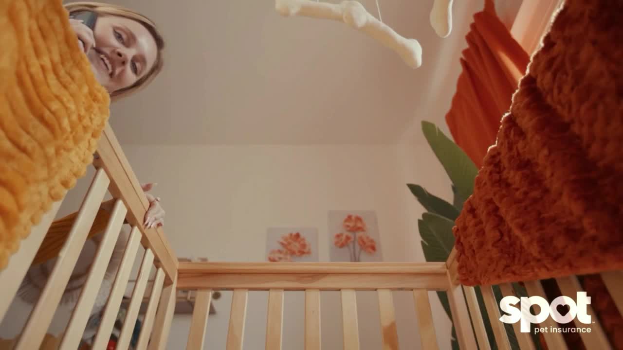 A woman leans over a crib, smiling while talking on a phone. A fluffy orange blanket frames the scene, with a mobile hanging above.
