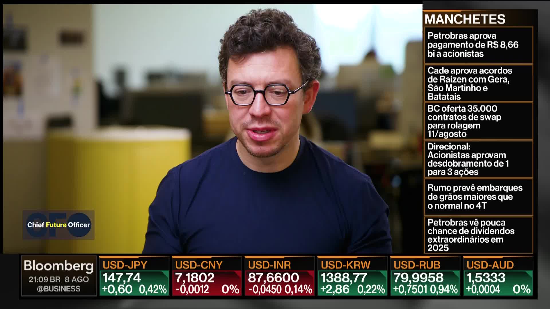 A man in glasses speaks directly to the camera on Bloomberg TV, with financial news headlines scrolling to his right. Below, currency exchange rates are displayed, with the time and the @BUSINESS Twitter handle visible.
