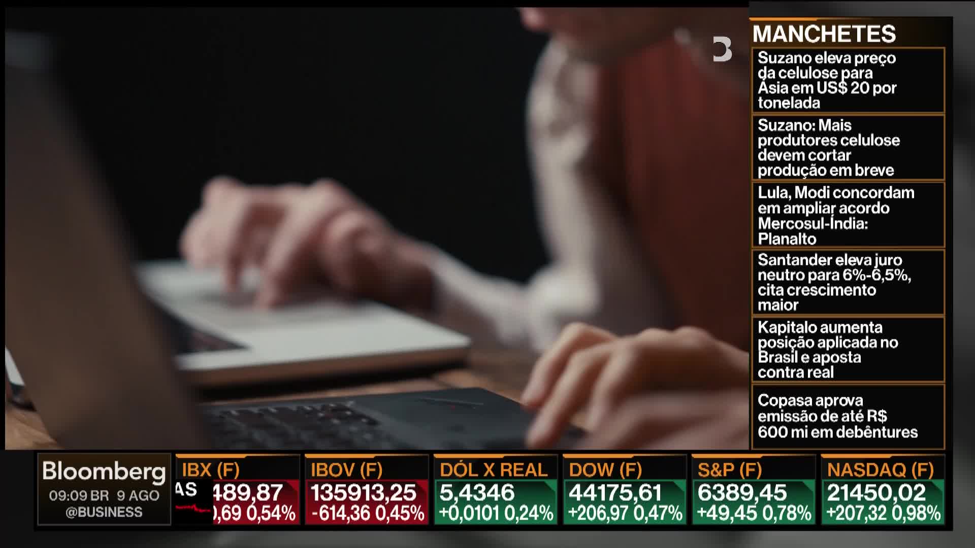 Someone's fingers tap across a laptop keyboard, their hands moving over the trackpad of another device. Below, Bloomberg TV's financial data scrolls across the screen.
