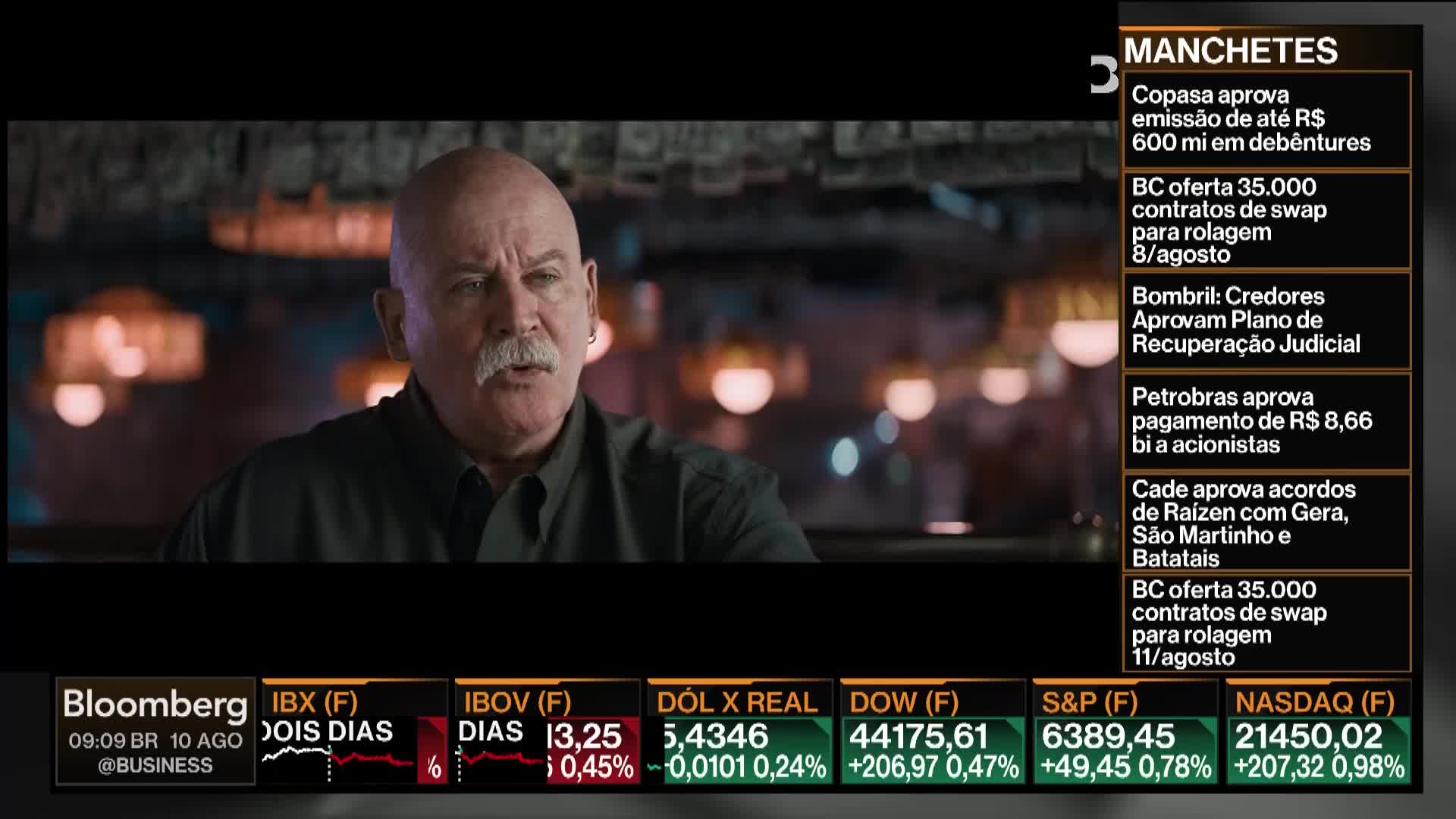A man with a white mustache speaks, lit by soft lights in the background. The Bloomberg TV ticker displays financial headlines and market data.
