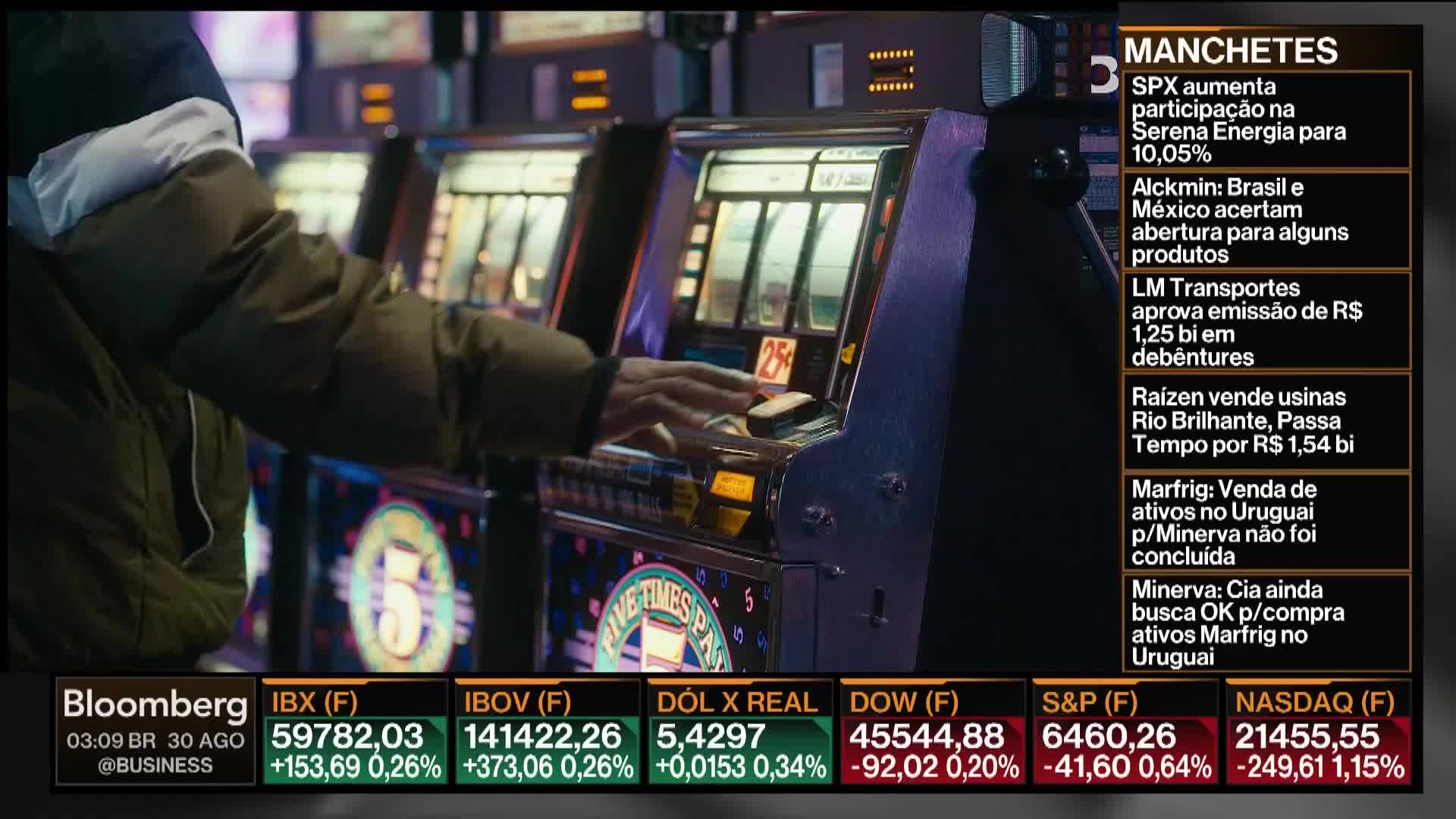 A person's hand rests on a slot machine, poised to pull the lever. The Bloomberg TV ticker displays financial news, with headlines in Portuguese.
