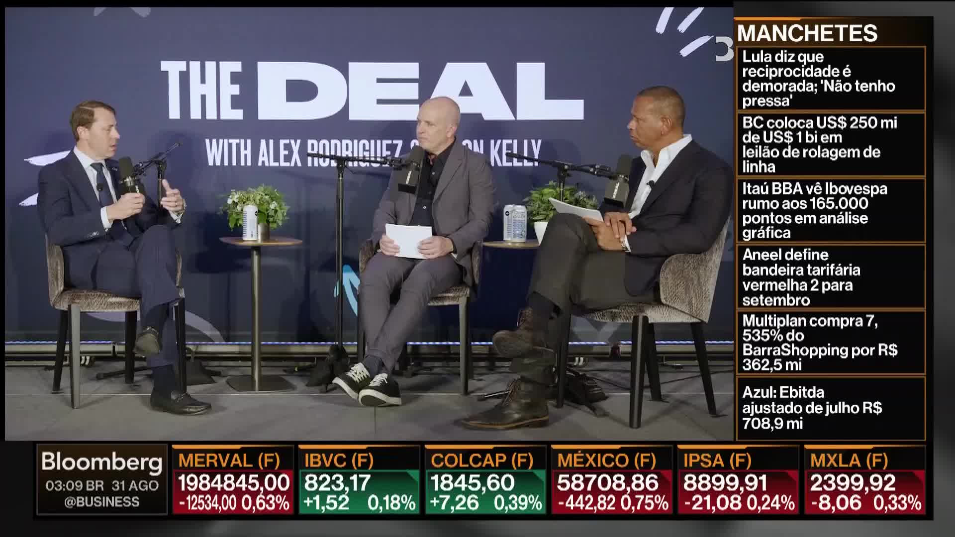 Three men sit on a set, microphones in front of them, talking. Bloomberg TV graphics run across the bottom and right side of the screen.
