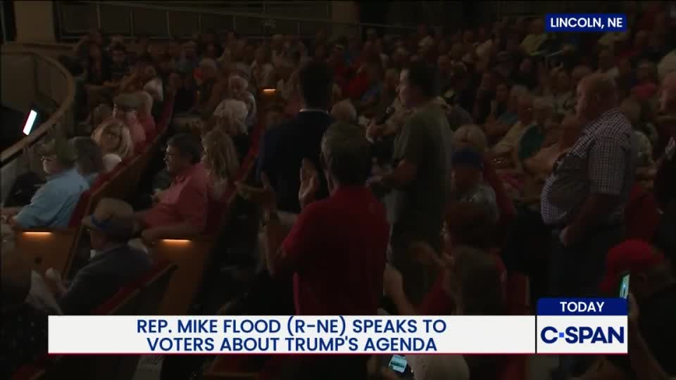 In a crowded auditorium, a man in a red shirt gestures as he speaks to someone off-camera. The audience, mostly older, sits in rows of seats, with C-SPAN cameras capturing the scene in Lincoln, Nebraska.
