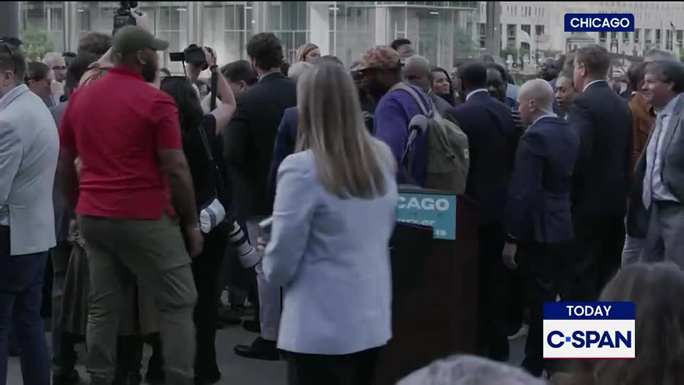 A crowd of people, some with cameras, surrounds a podium in Chicago. The C-SPAN logo is visible, indicating a live broadcast of the event.
