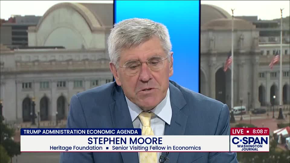 Stephen Moore, on C-SPAN, is speaking about the Trump Administration's economic agenda. Behind him, a backdrop shows a building with flags waving in front of it.
